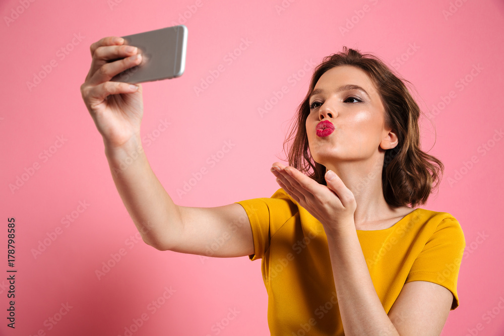 Poster close-up shot of young attractive woman with bright makeup sending air kiss while taking selfie on m