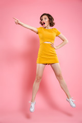 Full length portrait of shocked charming brunette woman with hand on her waist pointing with finger, looking aside while jumping over pink background