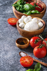 Ingredients for italian caprese salad. Mozzarella balls, buffalo, tomatoes, basil leaves, olive oil with vinegar, salt in olive wood bowls over gray texture background. Close up