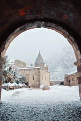 Armenian monastery Geghard, a view through an tuff arch entrance to a church yard, pavement and trees covered with snow with mountains and fog at the background.