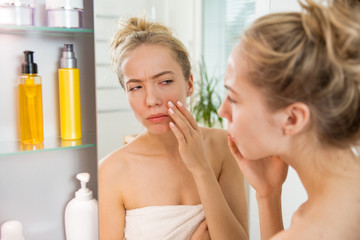 Young beautiful woman touching skin in bathroom. Unhappy girl standing in towel, looking in the mirror, checking dry irritated skin, puffiness and dark circles under eyes. Morning skincare routine.  