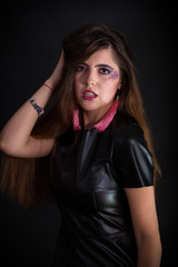 Indoors portrait of attractive young woman with makeup wearing black leather dress. Half-length standing and touching hair by arm. Dark background.