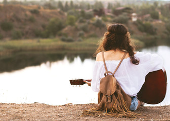 girl in boho style sits on a lake background with a guitar in hand