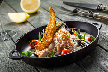 Delicious Asian meal made of sea bass fish, mussels, crab meat and other seafood with tomato and rice noodles on a rustic wooden table.