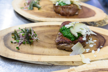 A meat fillet decorated with green pesto sauce and cheese shavings (pecorino nero) on a board made with a tree trunk portion accompanied by salad and green algae