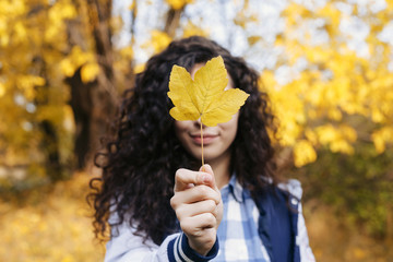 Beautiful young woman holding in hand one autumn yellow leaf. Girl closed face with maple leaf in hand. Outdoor. Holidays. Autumn.