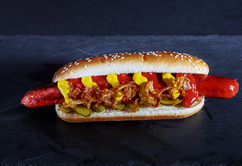 Delicious HotDog with a hot grilled sausage, fried bacon, salted cucumber, cheese sauce and ketchup, in a soft bun with sesame, on a dark stone background, fast food.