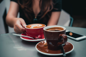 Closeup image of a woman's hand holding latte coffee cup with Americano coffee cup and smartphone on glass table in cafe