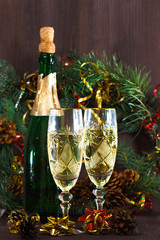 a bottle of champagne stands on a wooden background, next to two glasses and a wreath of fir branches with cones, it is hung with tinsel