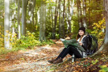 Beautiful brunette tourist girl wears black cap and backpacked have break with map and thermos of tea sitting near tree, autumn tourism concept