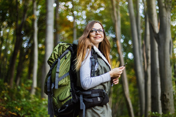 Cheerful brunette tourist backpacked girl have walk through forest holding smartphone, autumn tourism concept