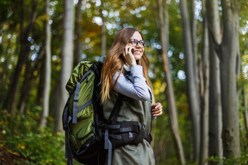 Fototapeta na wymiar Cheerful brunette tourist backpacked girl have walk through forest holding smartphone, autumn tourism concept
