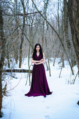 A young woman in an elegant burgundy evening dress in winter among trees and snow conceals a riddle