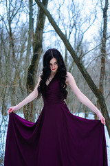 Fototapeta premium A young woman in an elegant burgundy evening dress in winter among trees and snow conceals a riddle