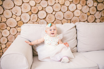 Fototapeta na wymiar little blue-eyed girl blond in a white tulle dress with a decoration on her head playing and rejoicing on a beige sofa in a room with decorative wooden wall in the form of hemp