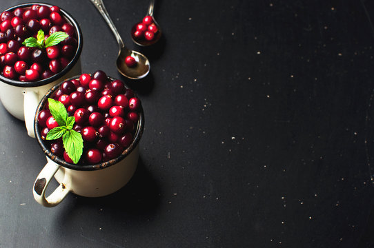Fresh red cranberries with mint leaves in a white cup and spoons on a dark black background with copy space.