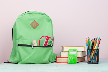Green backpack and school supplies: notepad, books, scissors, pens, pencils, ruler, calculator is on a white wooden table