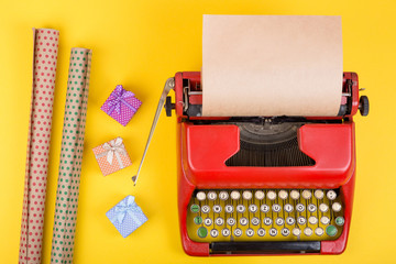 Holidays concept - red typewriter with blank craft paper, gift boxes on yellow background