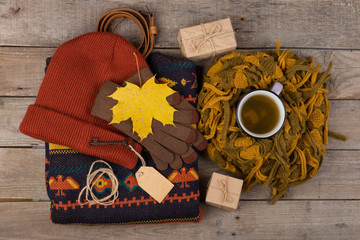 The concept of autumn coziness - a cup of tea, gift boxes, maple leaves, key with blank label, warm clothes: a hat, scarf and gloves on a brown wooden background