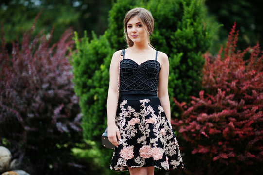 Portrait of a gorgeous young girl in black floral dress walking on the pavement with leather bag in the park on a prom day.