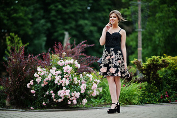 Portrait of a gorgeous young girl in black floral dress walking on the pavement with leather bag in the park on a prom day. © AS Photo Family