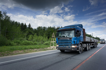 Blue truck driving on asphalt road in a rural landscape. Sunny summer day with a cloudy sky.
