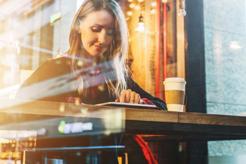 View through glass. Young smiling business woman sitting in cafe at table, drinking coffee and using tablet computer. Girl using digital gadget. Social network, online marketing, education,e-learning.