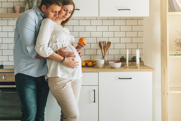 Young married couple embraces standing near table in kitchen. Husband hugs his pregnant wife, putting his hands on her big belly. A loving couple, future parents. Lifestyle, happy people.
