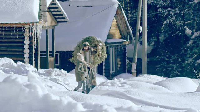 Lockdown of man with axe and woman in warm clothes carrying Christmas tree and walking along footpath in snow leading from wooden huts in winter forest; excited little boy running before them