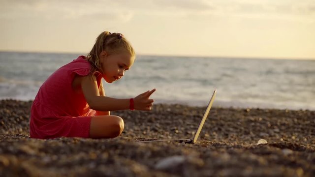 preschooler girl is sitting on a pebble beach in a sunset with laptop, pressing buttons