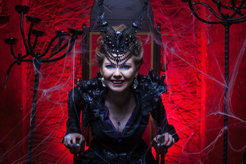 Woman in a black witch costume sits on a throne in a red room. She growls
