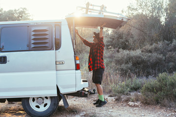 Urban adventurer and explorer stands next to his travel van or camping caravan in forest or field, during trip into wilderness with friends, opens truck of car in sunset light