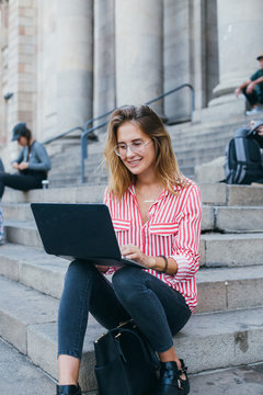 Beautiful and amazing blonde model in red shirt, with long straight hair, in trendy fashionable prescription glasses sits on stairs of office building or museum, works or study on laptop outdoors