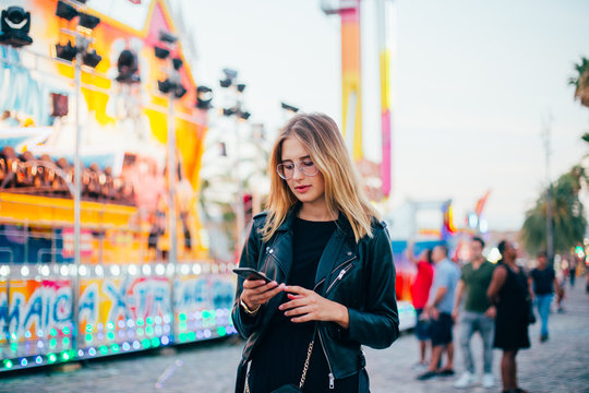 Trendy influencer modern millennial hipster teenager or cool fashionable woman stands in center of country fair or carnival or festival, uses smartphone app to stay connected on social media