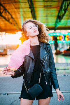 Fashionable young woman in black trendy leather jacket stands in middle of county fair, next to attraction rides or carousel, holds big pink cotton candy or sugar floss, enjoys date night or holidays