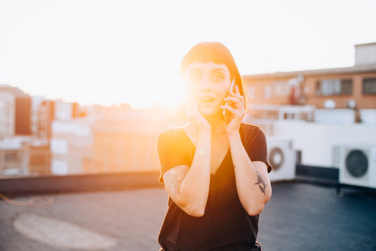Pretty young woman talks on phone and has excited or surprised face expression from news she just heard over smartphone call conversation from friend or partner, beautiful sunset light