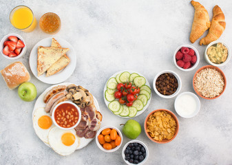 Fototapeta na wymiar Healthy varied breakfast, fried eggs, sausages, bacon and mushrooms with selection of fruits and vegetables, breads and juice on the grey white table, top view, copy space for text, selective focus