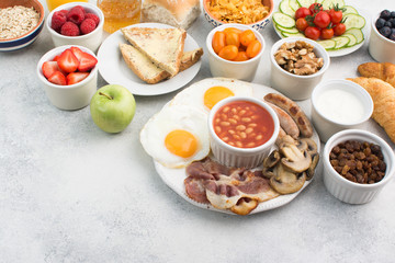 Fototapeta na wymiar English breakfast made with fried eggs, sausages, bacon and mushrooms with selection of fruits and vegetables, breads and juice on the grey white table, copy space for text, selective focus