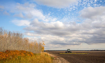 A tractor with an attached cultivator parked on a black dirt field surrounded by a forest in an autumn countryside landscape