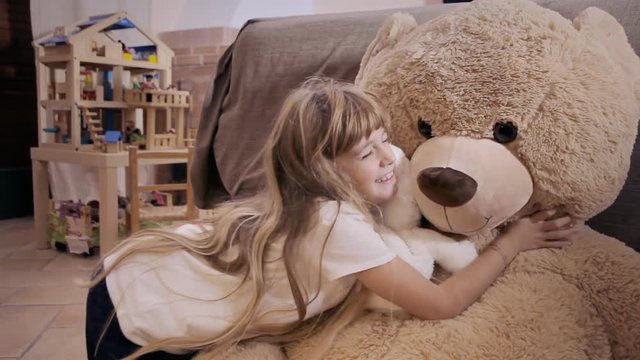 A cute little girl playing with her toy friends, a giant teddybear and a tiny one. The small bear says hi to the big one.
