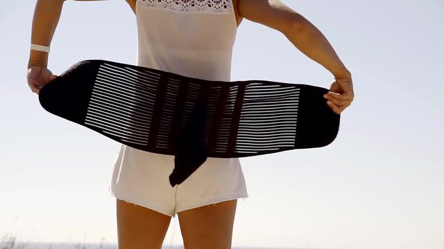 slim sporty woman is putting on remedial and supportive corset on a lumbar spine, standing outdoors