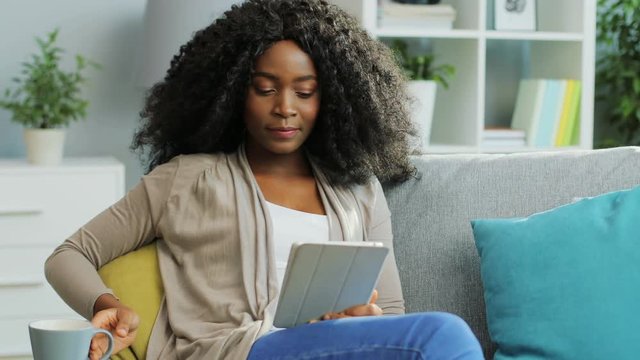 Curly African American young woman using her white tablet device and drinking tea. Indoor.