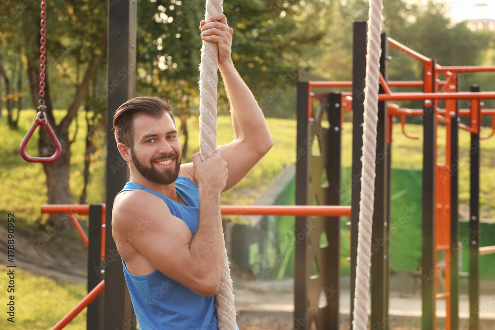 Wall mural Handsome young man doing rope climbing exercise outdoors