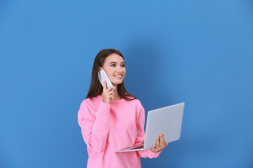 Young lady with laptop talking by phone against color wall