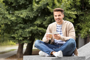 Obraz premium Young handsome man with cup of coffee using his mobile phone while sitting on parapet in park