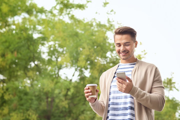 Young handsome man with cup of coffee using his mobile phone while walking in park