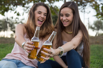 Two young laughing women posing on lawn in park and toasting with beer bottles having fun.  