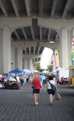 Marketplace under a bridge with booths setup and people shopping in the background with two women shopping walking in foreground shot from one end of the market during daylight