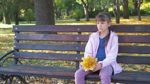 A sad little girl with yellow leaves sits on a bench.