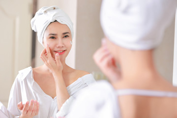 Young asia woman applying foundation or moisturizer on her face in front of mirror.